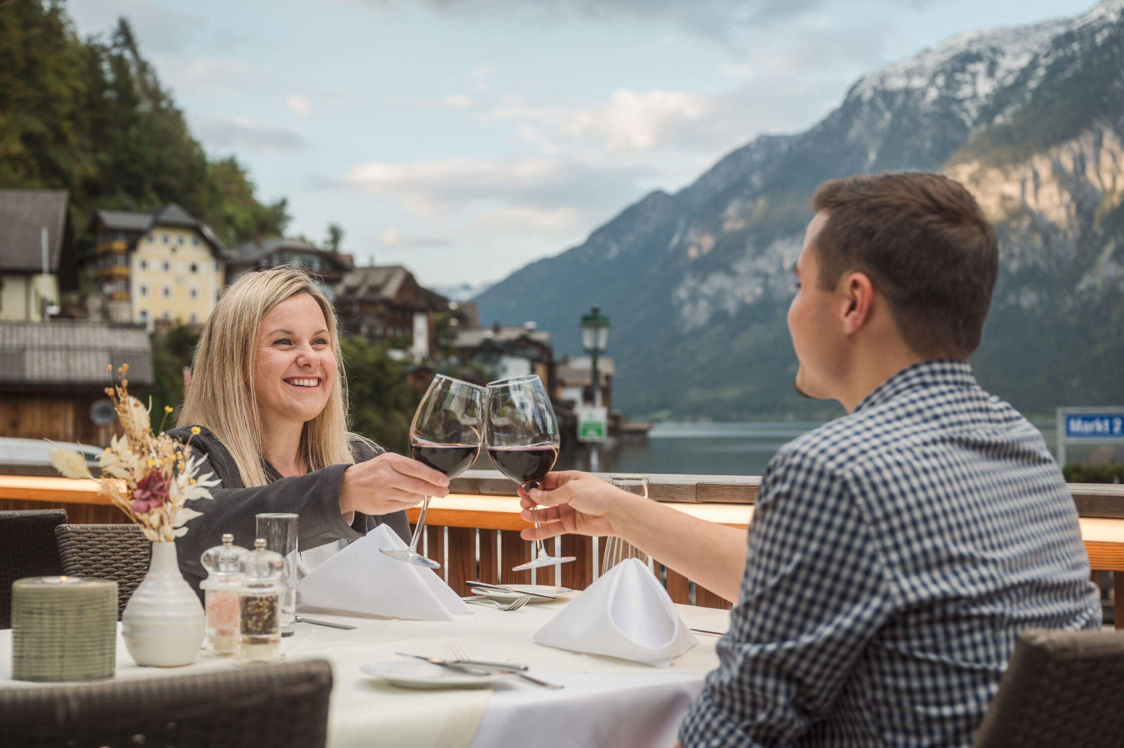 Ein Mann und eine Frau stoßen an einem Tisch mit Weingläsern an, im Hintergrund die Berge des Salzkammerguts.